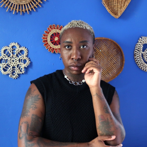 portrait of loveis wise against a deep blue background with round wicker sculptures. loveis is wearing a black muscle shirt and has tightly buzzed bleached hair. they have full sleeves of tattoos on their dark skin and are wearing a silver chain necklace.