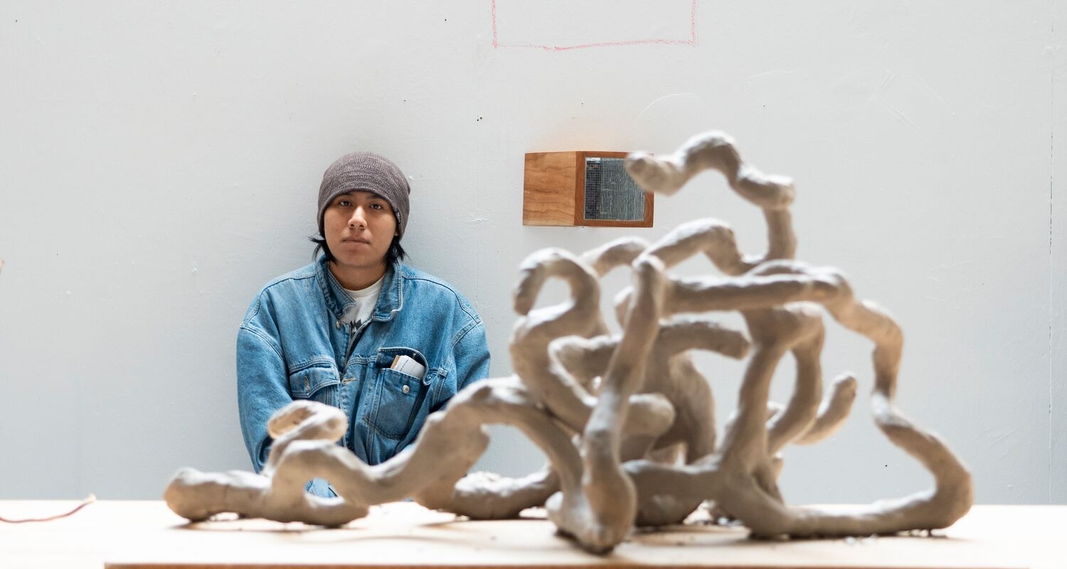 a student towards a back wall sitting square to the viewer looks across towards our right at a gnarly clay sculpture in the foreground resembling winding interlaced root or branch systems of a tree