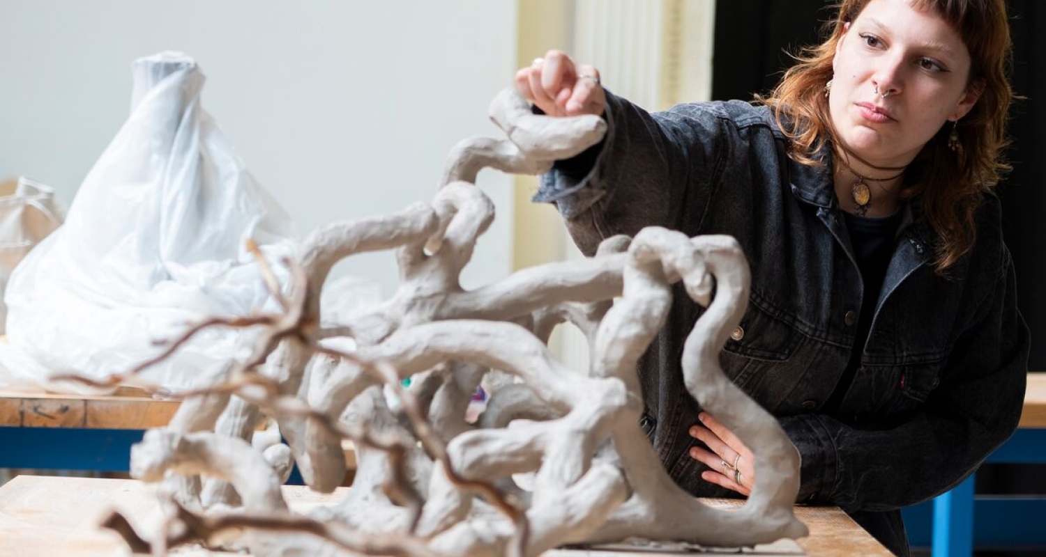 a person with golden-red hair reaches forward towards a gnarled clay sculpture resembling a larger snaking system of reference tree branches seen in the foreground. the person is pointing at the topmost point of the tangle