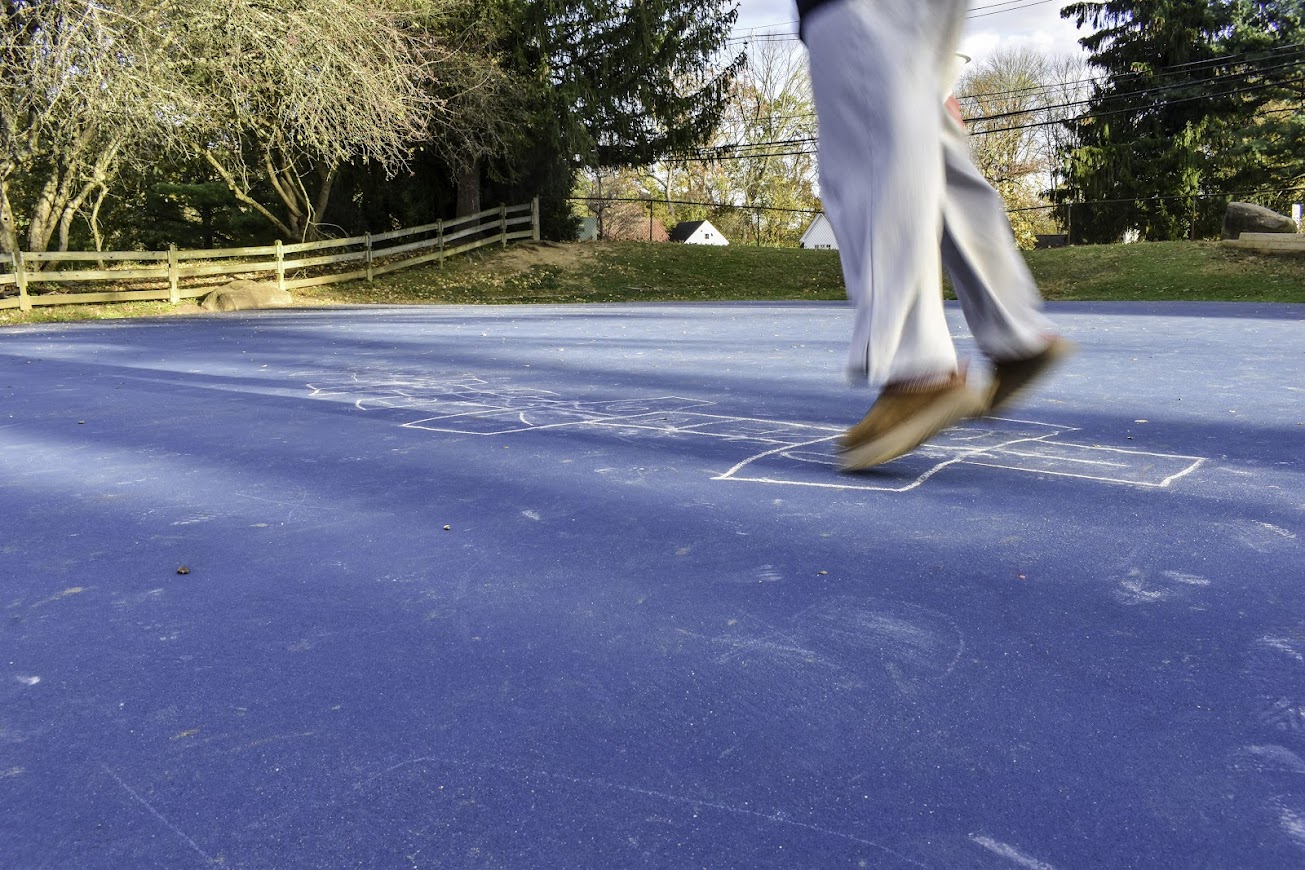 photograph of a person seen from the waist down wearing white pants and brown shoes playing hopscotch, with the grid shakily drawn with white chalk on an azure blue court, ringed by a wooden fence and green suburban scenery. the person's shape has some motion blur. 