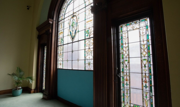Interior shot of a beautiful set of stained glass windows surrounded by detailed ornate dark wood trim. The windows have three panels, the outer two are rectangular and roughly door sized, reaching up from the pale green floor. The central window is massive and is vaulted. 