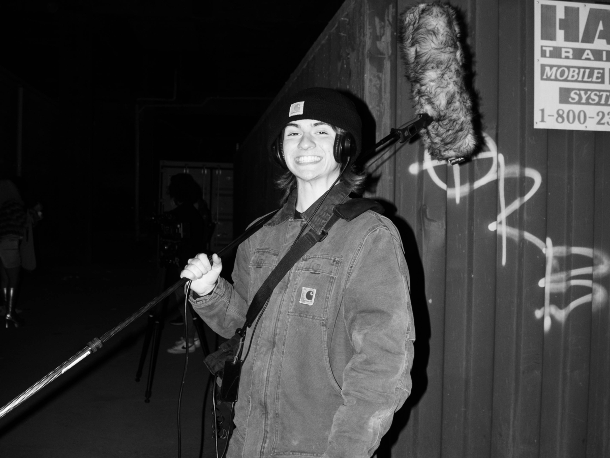 a black and white image of a person holding a boom mike on their shoulder and smiling at the viewer against a background of a metal shipping container sprayed with tags. 