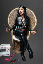 kristina wong in a black leather outfit including a cap sits in a high-back wicker throne holding fabric working supplies. next to her tall black riding boots is a hello ktity branded sewing machine and sewing supplies