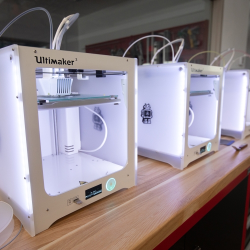 A row of glowing 3D printers sit on top of a wooden table in UArts' Makerspace