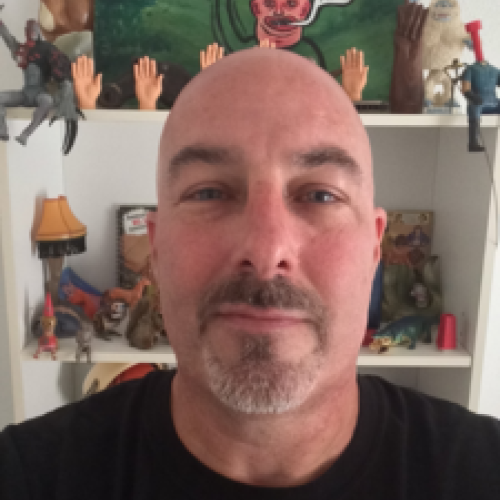 John Serpentelli head shot, bald, black t shirt, in front of bookshelf with small plastic hands and a cartoon painting