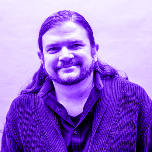 headshot of james cowen overlaid with violet. james is wearing a thick cardigan over a collar shirt and has long hair brushed back from a bearded face. 