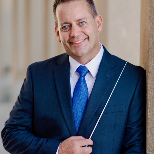 Keith Hodgson in a blue suit with a blue tie and white shirt holding a conductors baton