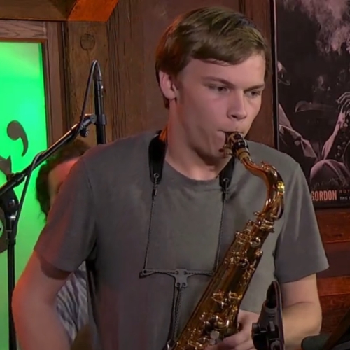 photo of matt miller playing the saxophone, focused and looking at notation to the right outside of the frame of the photo. matt is wearing a gray-green shirt and has wavy golden hair and is wearing an apply watch