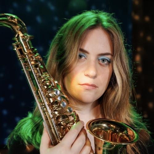 headshot of kaitlyn oliveri holding a shining saxophone up. kaitlyn has long blond hair, lit up with a green spotlight from above. speckled trippy lighting marks the dark background. kaitlyn has strong brows and pale blue eyeshadow