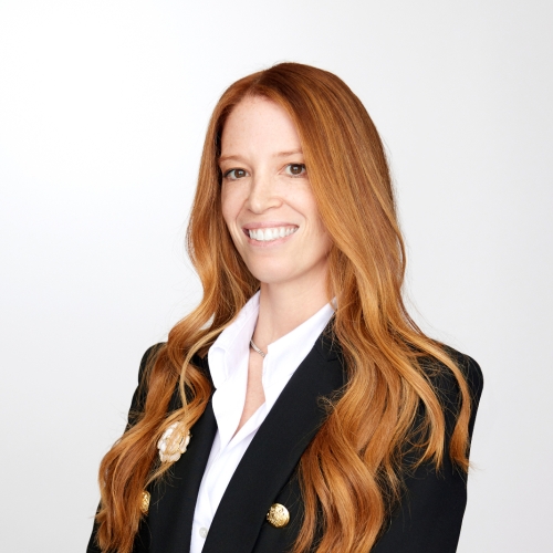 headshot of rachel pritzker against a plain white background wearing a black blazer with gold buttons. rachel has very long flowing red gold hair. 