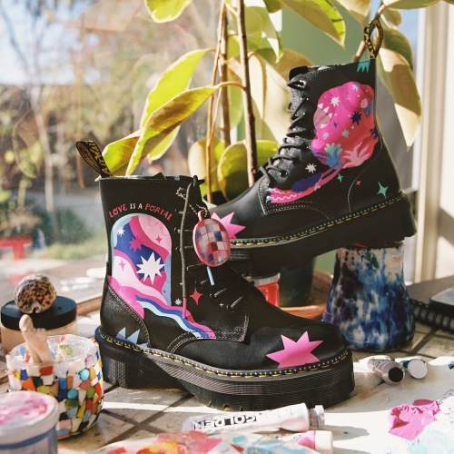 a pair of doc martens high-top boots with fantastical illustrations of cosmic gateways, stars, and sparkles rendered in pastel pinks, vivid fuchsias, blues and cyans. the shoes are resting in a sunny studio space among paint tubes.