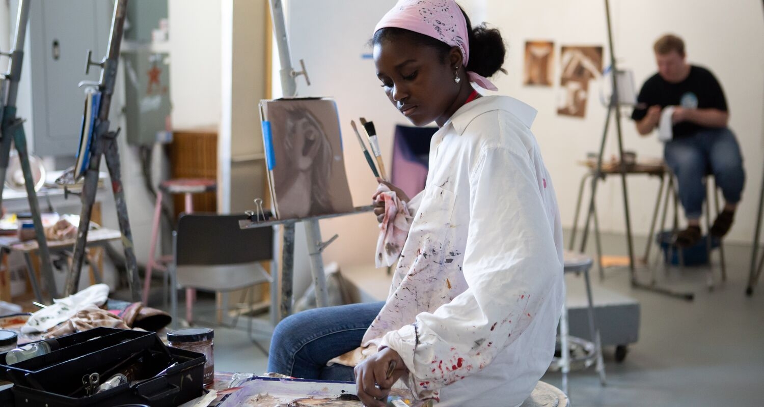 a student in a white paint-splatter shirt sits at an easel and reaches over to get more paint. another student sits at an easel in the background. 