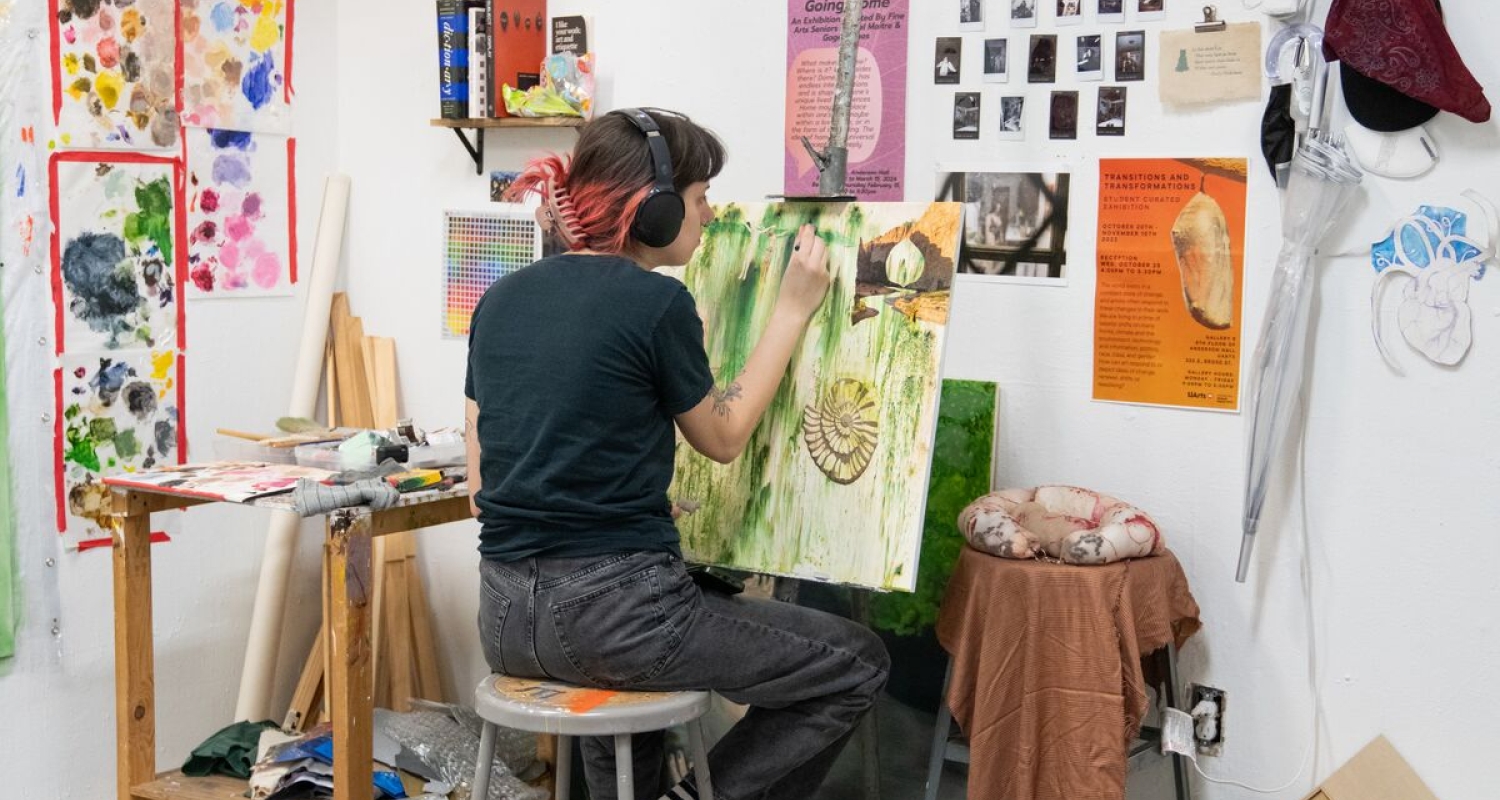 a student seen from behind working on a painting on an easel in a white-walled studio corner covered in test splotches of paint and inspiration imagery. 