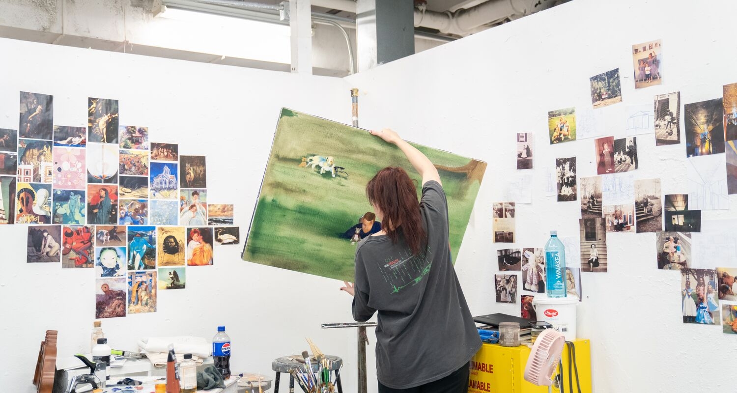 a student handles a medium-large canvas onto an easel. it depichs a pale green pastoral scene. the walls of the white studio corner are covered in a tightly arranged grid of many many inspiration iamges