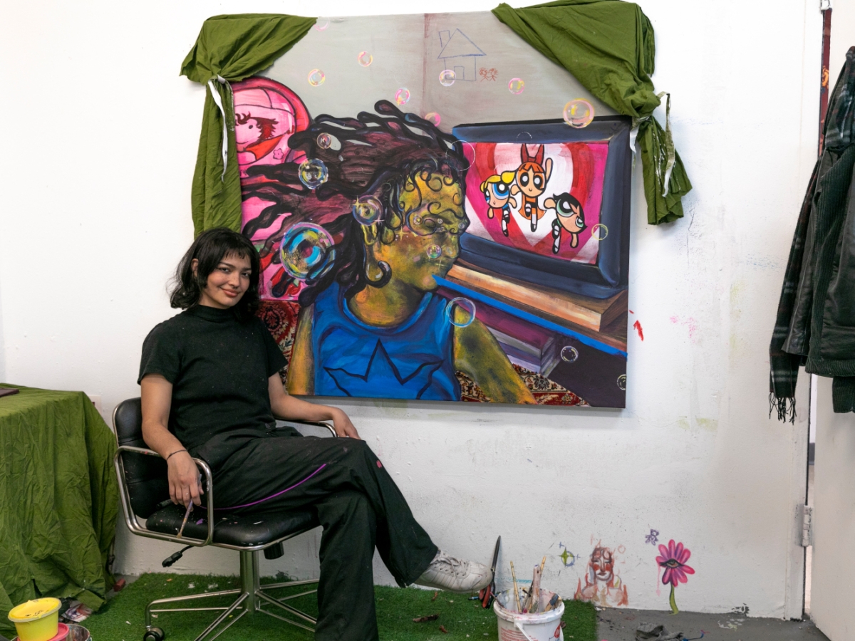 a fine arts student sits at their canvas depicting a person with a molten face in a blue top and a power puff girls episode in the background. the painting is surrounded by green drapes. 
