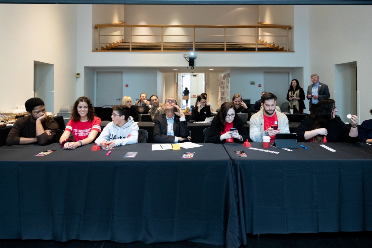 interior of CBS auditorium during the day of giving event. several rows of black cloth-drape desks are in the room with staff and mostly red t shirts attending to matters on phoen and tablets. above the tables is the upper balcony seating area of the auditorium. double doors are seen directly behind the desks in the next room leading out into sunlight.