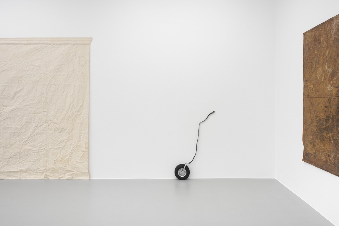 Installation view of small tire with leash attached to wall and two other cloth works in frame