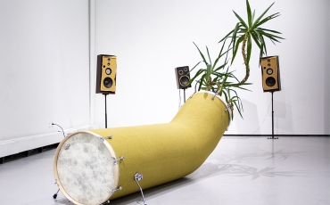 An installation comprising a long drum fashioned into a tube with a tree coming out of the tube and surrounded by speakers.