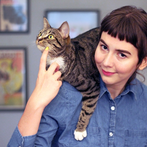 Jameela Wahlgren in a denim button down shirt with a gray cat on her shoulder that she is pointing to while looking at the camera