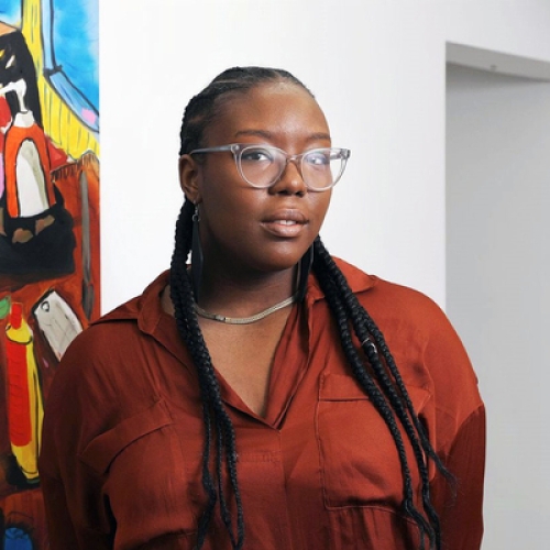 Patricia Renee Thomas wearing a brown blouse standing in front of a white wall with the edge of a painting on one border 