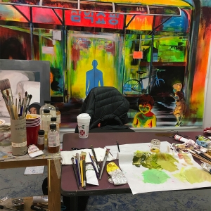 A student's painting studio. Paints, brushes, paper towels and other supplies are laid across the table with a canvas hanging on the wall behind.