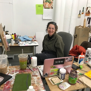 A student sits in their painting studio behind their computer and smiles at the camera.