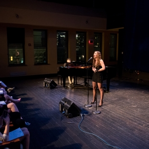 a person in a short black dress sings into a microphone on a warmly illuminated stage as a grand piano accompaniment is seen directly behind. audience members poke into the shot from the left, and a nighttime cityscape is seen beyond the dark windows. 