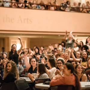 interior of levitt auditorium during new student orientation. the view is approximately from in front of the stage, looking into a thick audience. all of the raked auditorium seating is filled, with additional audience members seated in the foreground at folding tables and chairs, along with people in the upper autidorium balcony. the crowd is smiling and cheering, with a person at the left of the image standing with a hand raise, yelling. 