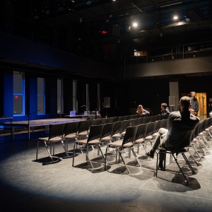 interior shot of caplan center's black box theater seen from left of the seating, which is in flat rows on the floor right in front of a stage at the left of the image. the stage is pressed up against thing tall windows. 