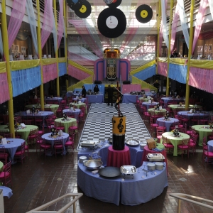 solmssen court decorate for a music-themed event in pastel colors. giant faux vinyl records hang from the ceiling as the cross stairwells across from CBS auditorium are decorated to resemble a jukebox. dining tables are in pastel violet, green colors with red and pink accents, as long streamers and drapes in blue, pink, and white cover the space. 